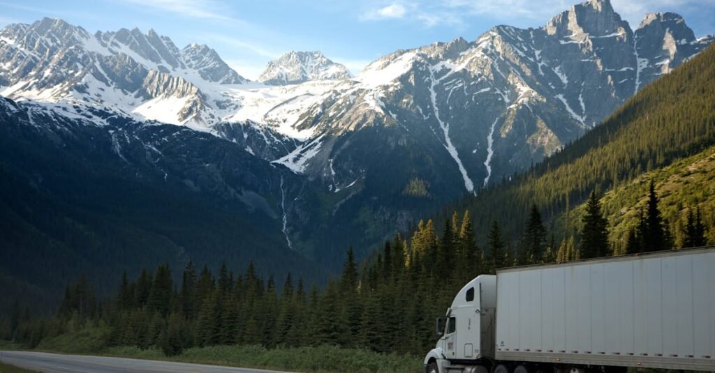 A semi-truck travels along a highway with snow-capped mountains in the background.